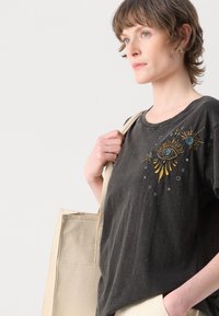 T-shirt in cotone grigio scuro con ricamo di sole e occhio in oro e blu. Il modello tiene in mano una borsa a tracolla beige, mettendo in evidenza la vestibilità morbida della maglietta.