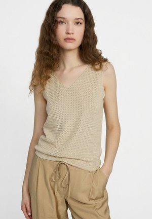 Frau mit welligem braunem Haar, die ein beiges ärmelloses Strickoberteil und lockere beige Hose mit Kordelzug trägt, vor einem einfachen weißen Hintergrund.