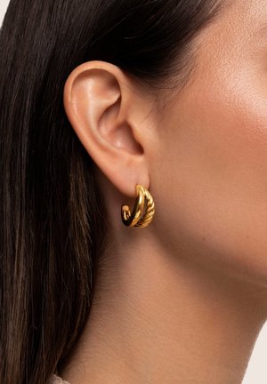 Gros plan sur l'oreille d'une femme portant une boucle d'oreille en or torsadé, avec des cheveux foncés et une peau lisse visibles.