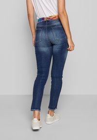 Smala blå jeans med fransad nederkant, dekorerade med ett färgglatt mönstrat midjeband och subtila utsmyckningar på bakfickorna.