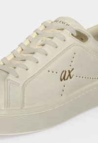 Gros plan sur une basket beige avec lacets, étoiles perforées et les lettres "ax" sur le côté en marron.