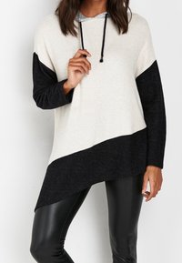 Kvinna som bär en asymmetrisk svart och vit långärmad hoodie i kombination med svarta läderleggings, stående mot en enkel vägg.