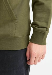 Olive green sweatshirt featuring a textured surface, ribbed cuffs, kangaroo pocket, and brand label on the side, paired with dark jeans.