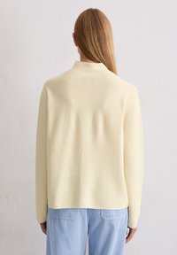 Kremowy sweter dzianinowy z kołnierzem typu mock neck, długimi rękawami i luźnym krojem, w połączeniu z jasnoniebieskimi spodniami w prążki. Widoczne detale teksturowane.