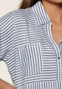 Camisa de cuello con rayas verticales en azul y blanco, que presenta un bolsillo en el pecho y cierre frontal de botones, hecha de una tela ligera.