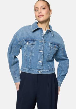 Frau mit heller Haut und zurückgebundenen Haaren, die eine blaue Jeansjacke mit geknöpften Pattentaschen und dunkle, hoch taillierte Hosen trägt.