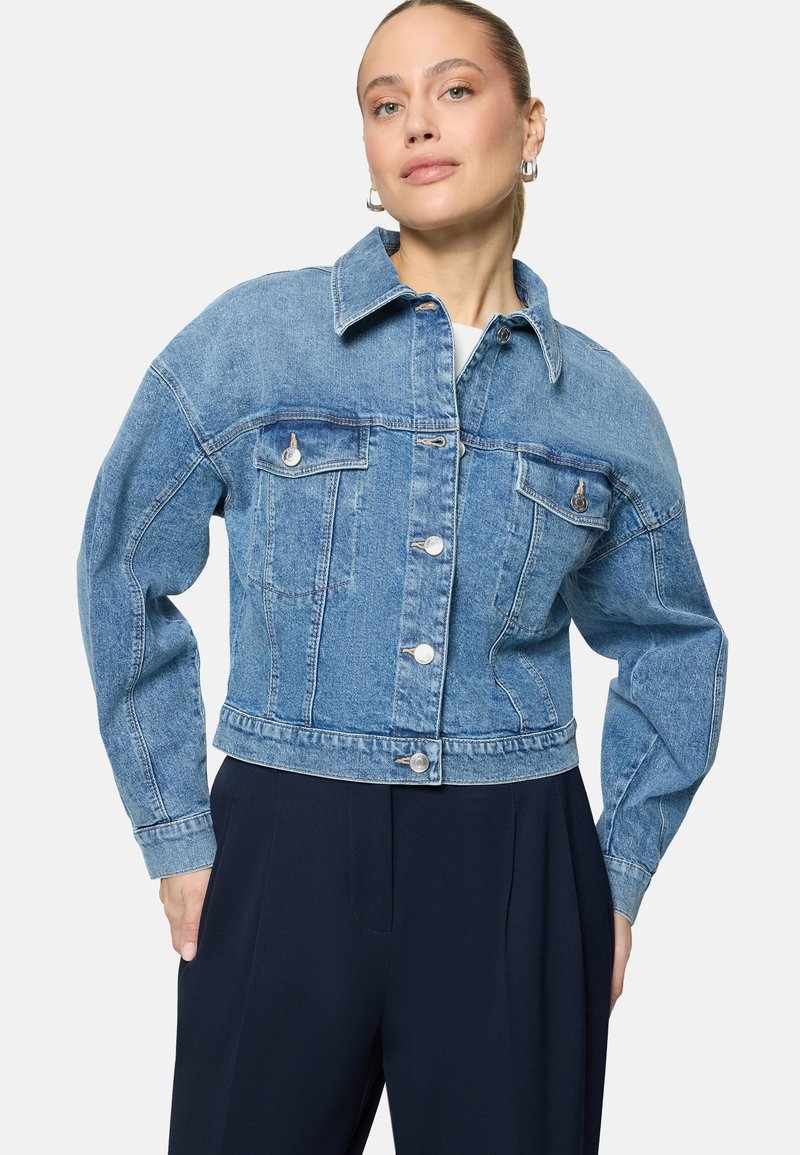Femme à la peau claire et aux cheveux attachés, portant une veste en denim bleue avec des poches à rabat boutonnées et un pantalon foncé taille haute.