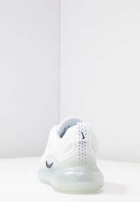 Baskets blanches Nike avec semelle translucide et petits points foncés, vues de dos sur une surface blanche et un fond uni.