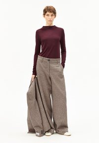 Bordeaux lange mouwtop en brede bruine tweedbroek met een knoopsluiting. Het model houdt een bijpassende bruine tweedjas vast. Lichte achtergrond.