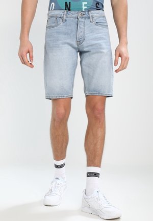Homme portant un short en jean bleu clair, des chaussettes blanches avec une rayure noire et des baskets blanches, debout devant un fond uni.