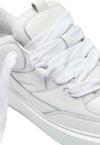 Valkoiset tennarit, joissa on teksturoitu nahkapinta, paksut valkoiset nauhat, pehmustettu kieli ja sileä kumipohja. Minimalistinen muotoilu, jossa on hienovaraisia yksityiskohtia.