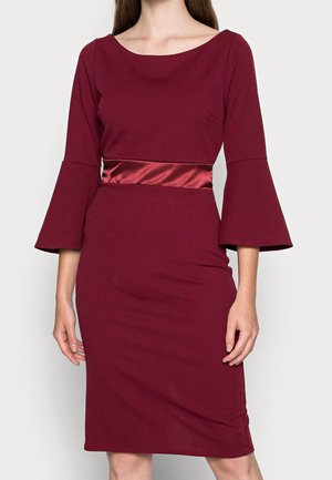 Woman wearing a fitted burgundy knee-length dress with bell sleeves and a shiny satin waistband.