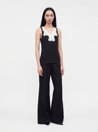 Femme aux longs cheveux foncés portant un haut sans manches noir avec des détails en dentelle blanche et un pantalon noir à jambes larges, debout devant un fond uni.