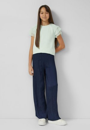 Jasnozielony top z warstwowymi falbanami na rękawach, zestawiony z granatowymi plisowanymi spodniami o szerokich nogawkach i białymi trampkami. Gładkie tekstury, luźny styl.