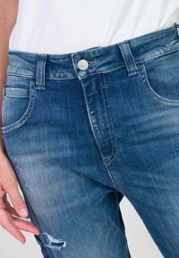 Jean en denim bleu avec une coupe ajustée, présentant une fermeture à bouton en métal, un léger effet usé sur la cuisse gauche et un style classique à cinq poches.