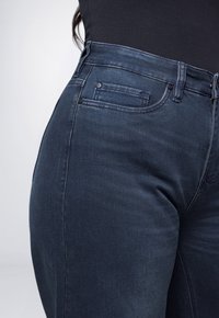 Persoon in donkerblauwe denim jeans en een zwart top, waarbij de taille en het bovenbeen zichtbaar zijn met zichtbare stiksels en zakken.