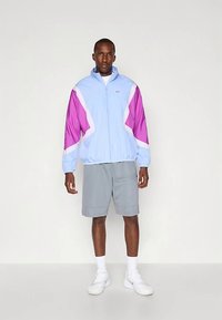 Casaco corta-vento Nike em azul claro com detalhes em rosa e branco, apresenta fecho de correr frontal, punhos elásticos e ajuste descontraído, combinado com calções cinzentos.