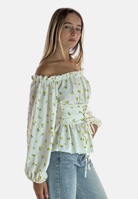 Weiße schulterfreie Bluse mit kleinem gelben Blumenmuster, gerafftem Ausschnitt und gerafftem Taillenbereich; kombiniert mit hellblauen Jeans.