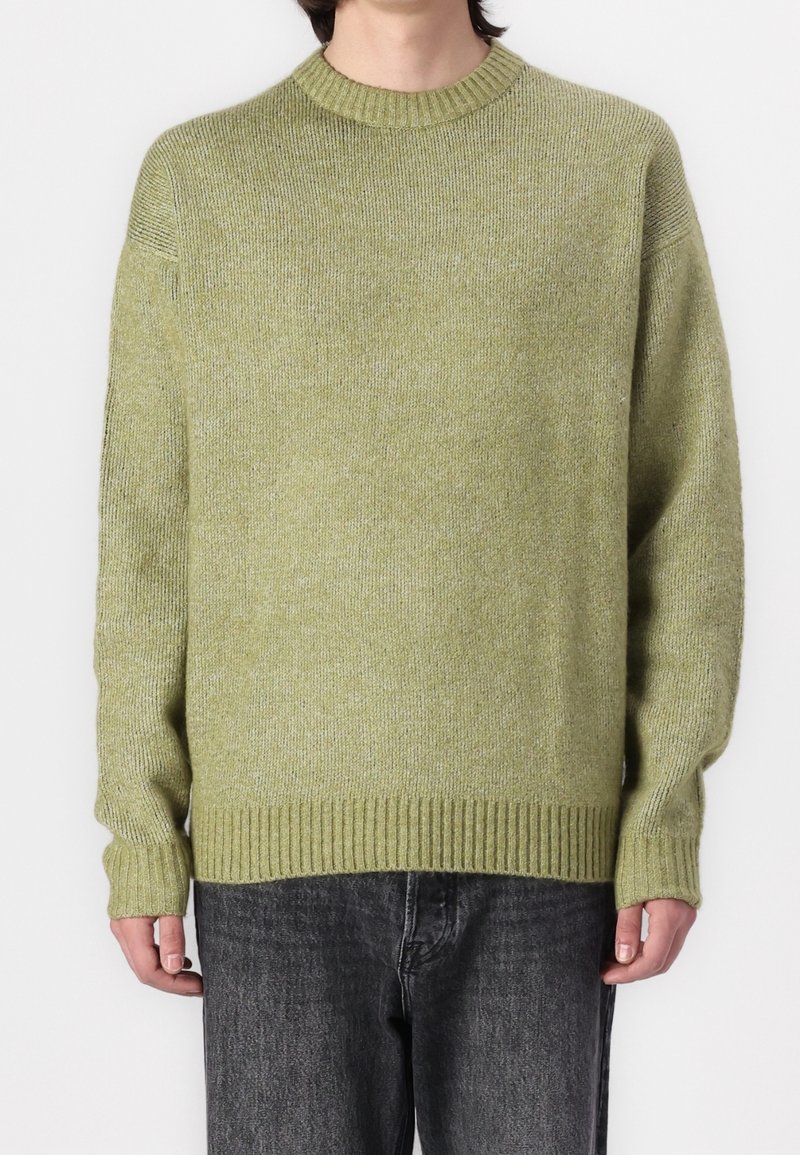Jasnozielony, oversize’owy sweter z dzianiny z prążkowanym dekoltem i dolną krawędzią. Charakteryzuje się teksturowaną tkaniną i opuszczonymi ramionami. Noszony na ciemnych jeansach.