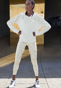 Weiße Kapuzenjacke mit goldenen Wirbelmustern, kombiniert mit cremefarbenen Leggings, die ähnliche Wirbel aufweisen; das Modell trägt schwarz-weiße Sportschuhe.