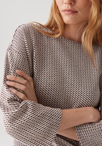 Femme aux cheveux châtain clair croisant les bras, portant un haut à manches longues texturé beige et noir avec un motif diagonal.