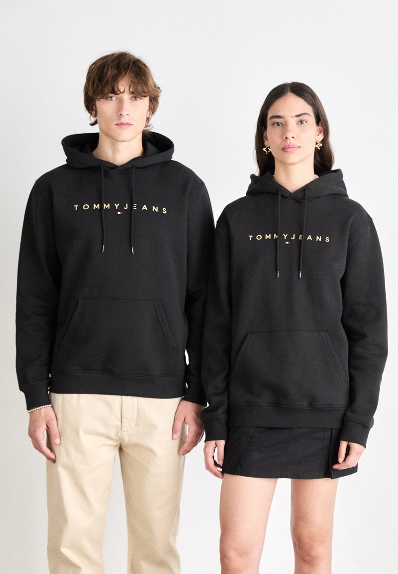 Sudaderas negras con el texto dorado "TOMMY JEANS", bolsillos tipo canguro y cordones. Un modelo lleva pantalones beige y el otro una falda negra.