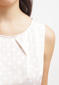 Ljust rosa ärmlös topp med vita prickar, med en plisserad halsringning och slät tygkvalitet.