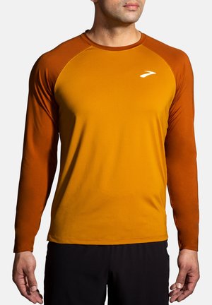 Mann trägt ein langärmliges Laufshirt in Senfgelb mit brennend orangefarbenen Ärmeln und einem weißen Logo auf der Brust.