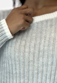 Maglioncino grigio chiaro a maglia con trama a coste verticali, dotato di un colletto e polsini bianchi. Una mano sta afferrando il collo.