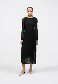 Sieviete stāv, valkājot melnu midi kleitu ar teksturētām caurspīdīgām garām piedurknēm un svārkiem, valkājot baltas smailas kurpes ar siksniņu pie papēža.