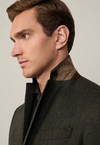 Cappotto a spina di pesce verde scuro con colletto alto e interno tessuto marrone strutturato. Stile a taglio corto che mette in risalto il colletto e il materiale.