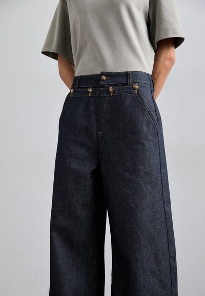 Pantalon large en denim bleu foncé avec poches avant, boutons dorés et surpiqûres contrastées. Associé à un haut gris clair à manches courtes.