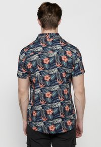 Korte mouwen shirt in marineblauw met een tropisch bloemenpatroon featuring roze hibiscus en groene bladeren. De stof lijkt lichtgewicht en ademend.