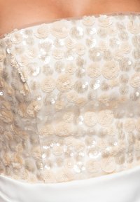 Gros plan sur le corsage d'une robe blanche sans bretelles ornée de sequins floraux beiges et de délicats embellissements superposés.