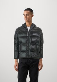 Donkergroene pufferjack met een glanzende nylon stof. Voorzien van een capuchon, een rits aan de voorkant en twee zijzakken. Iets oversized pasvorm met een gestikte ontwerp.
