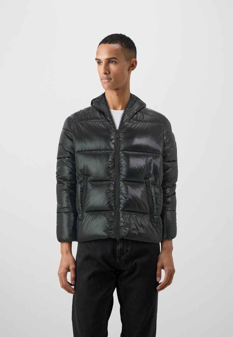 Donkergroene pufferjack met een glanzende nylon stof. Voorzien van een capuchon, een rits aan de voorkant en twee zijzakken. Iets oversized pasvorm met een gestikte ontwerp.
