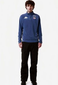 Maglione in pile blu con collo a mezza zip, con un logo bianco sul petto. Abbinato a pantaloni cargo neri e calzature nere.