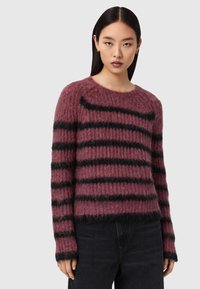 Jeune femme portant un pull duveteux mauve avec des rayures horizontales noires et un pantalon noir, sur un fond gris uni.