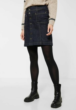 Vrouw draagt een donkerblauwe spijkerrok met knopen aan de voorkant, zwarte ondoorzichtige panty's, zwarte enkellaarsjes en een beige trui met patroon aan de manchetten.