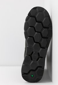 Semelle de chaussure noire avec motif de bande de roulement hexagonal et un petit hexagone vert près du talon, positionnée verticalement sur un fond blanc.