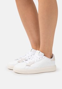 White athletic shoes with perforated accents, a smooth leather surface, beige details, and a textured rubber sole. Laced design and padded tongue.