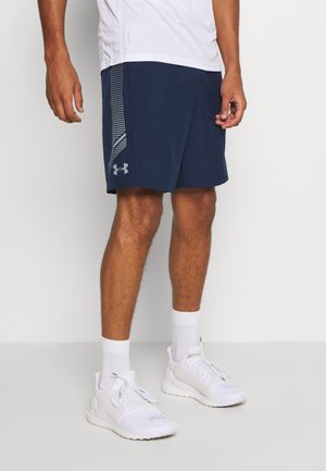 Man draagt marineblauwe Under Armour shorts met een wit patroon op het zijpaneel, witte sportschoenen, witte sokken en een wit shirt zichtbaar bij de taille.