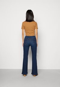 Brun kortärmad topp ihop med hög midja mörkblå utsvängda jeans. Märkbar söm på bakfickorna. Modell vänd bort från kameran.