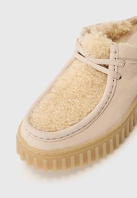 Beige slip-on shoes featuring a fluffy cream lining, textured outsole, and subtle stitching details. Soft material with rounded shape.