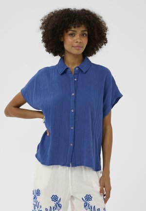 Žena s kučeravými vlasmi, oblečená v modrej textúrovanej košeli s krátkym rukávom na gombíky a bielych nohaviciach s modrou kvetinovou výšivkou, ruka na boku.