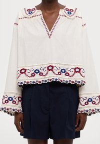Blusa blanca con patrones florales bordados en rojo y azul, con escote en V y mangas anchas con un acabado decorativo. Combinada con pantalones cortos azul marino.