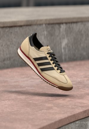 Beige Adidas sneaker met zwarte strepen en veters zwevend boven een roze betonnen oppervlakte buiten.