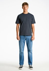 Laivansinis puuvillainen t-paita, jossa on pyöreä kaula-aukko, yhdistettynä keskipitkiin sinisiin farkkuihin ja musta-valkoisiin tennareihin mallin päällä valkoista taustaa vasten.