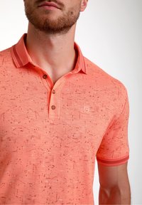 Trumpas rankovėmis oranžinis polo marškinėlis su tekstūruotu raštu, dviem mygtukais, kontrastingu apykakle ir logotipu krūtinėje.