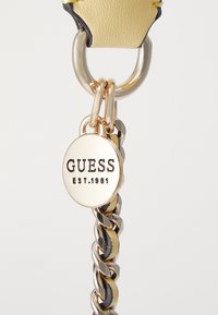 Llavero de imitación de cuero amarillo con una cadena de metal dorado y una etiqueta circular grabada con "GUESS EST.1981". Textura brillante y suave.